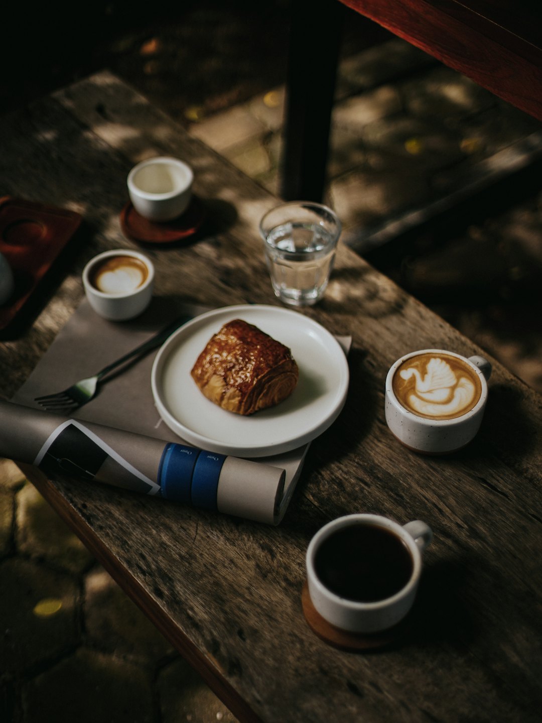 Mesa de desayuno con tazas de café, un cruasán y un periódico.